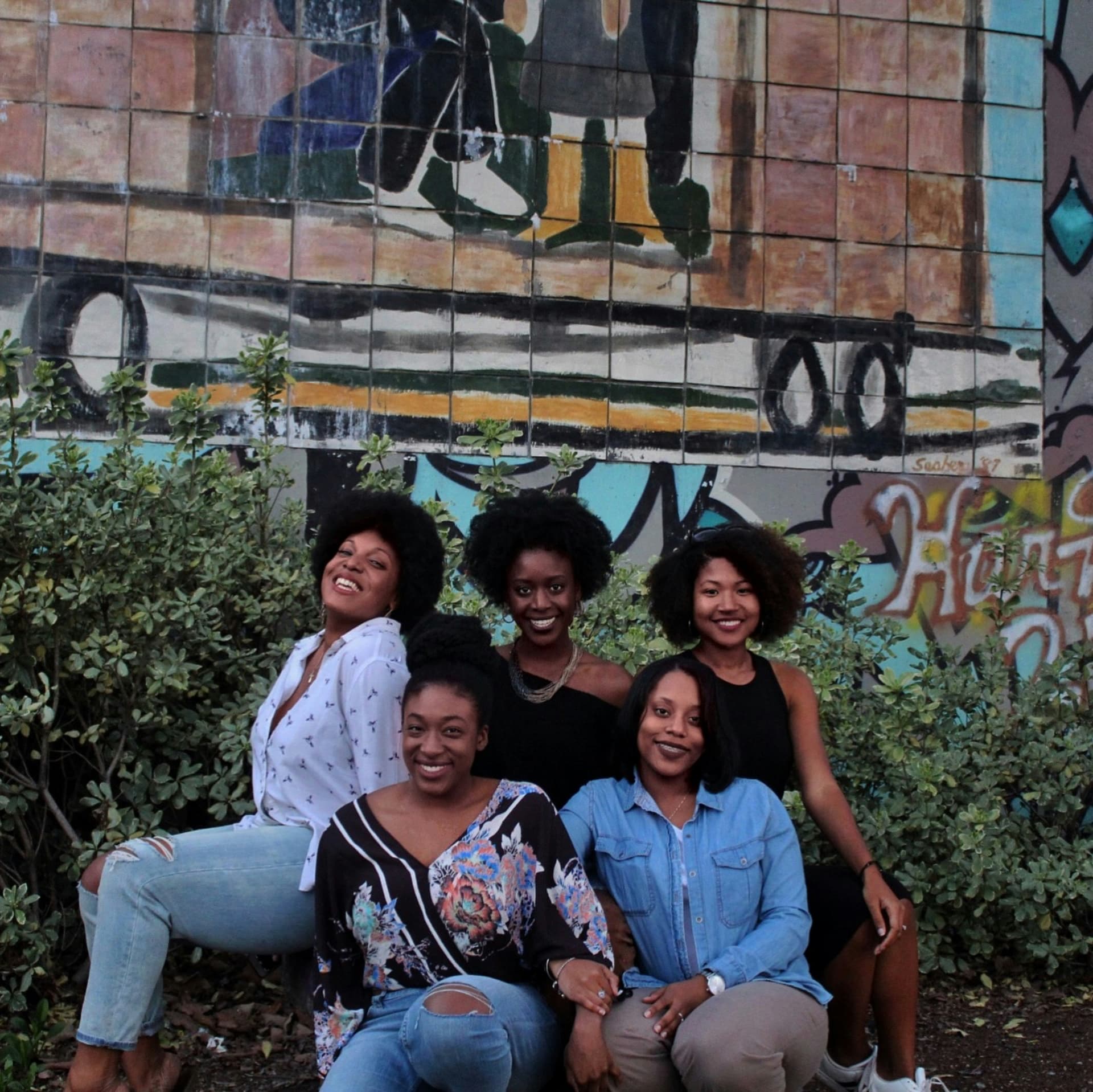 Five women smiling together in front of a colorful tiled mural wall